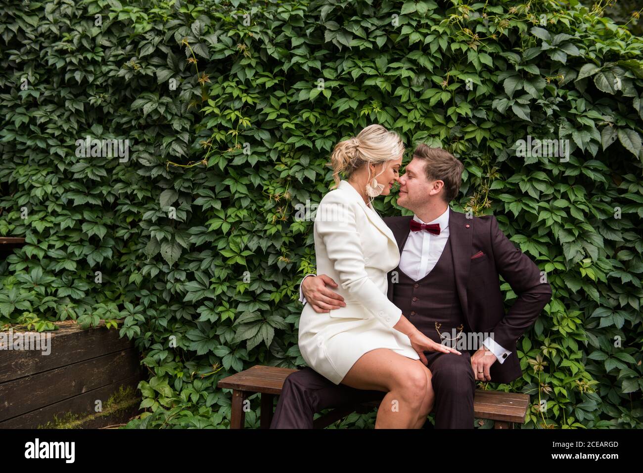 Uomo e donna eccitato indossando abiti da sposa alla moda e guardando l'un l'altro contro la siepe verde Foto Stock