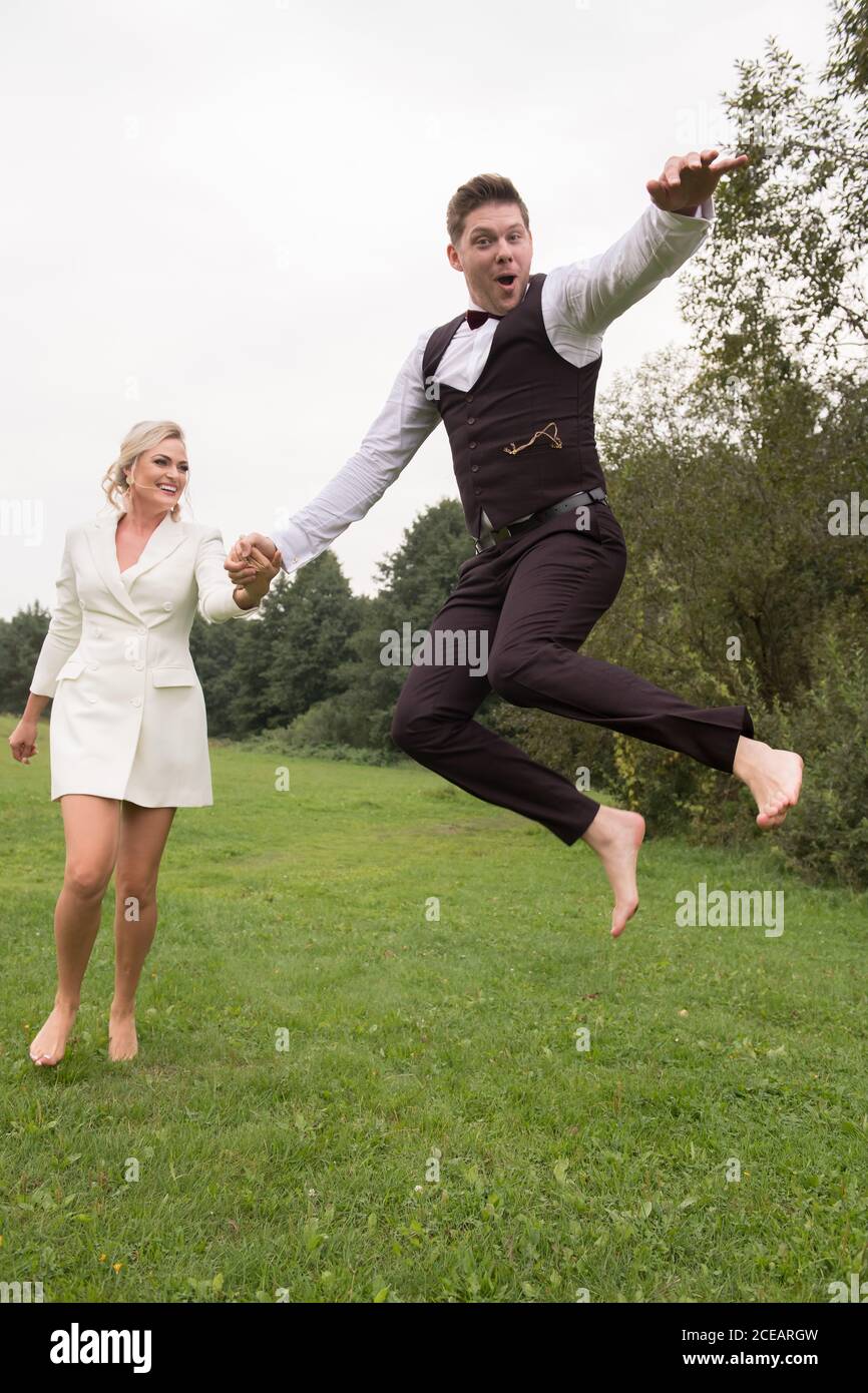 Sposo e sposa adulti alla moda in abiti eleganti che tengono le mani e saltando con l'eccitazione sul prato verde Foto Stock