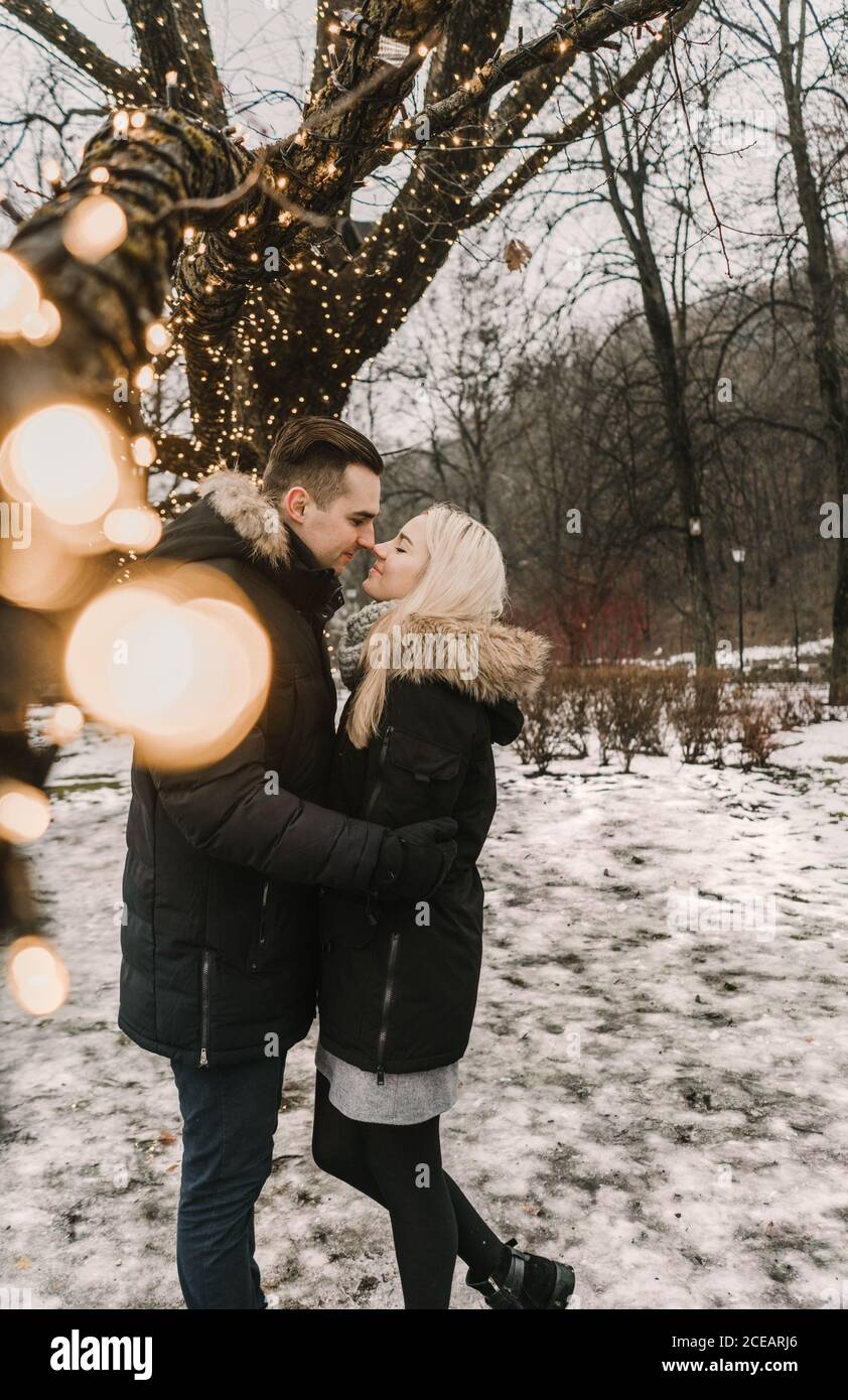 Vista laterale di felice guy kissing young lady in giacche sci nei pressi di legno decorate da accese le luci fairy Foto Stock