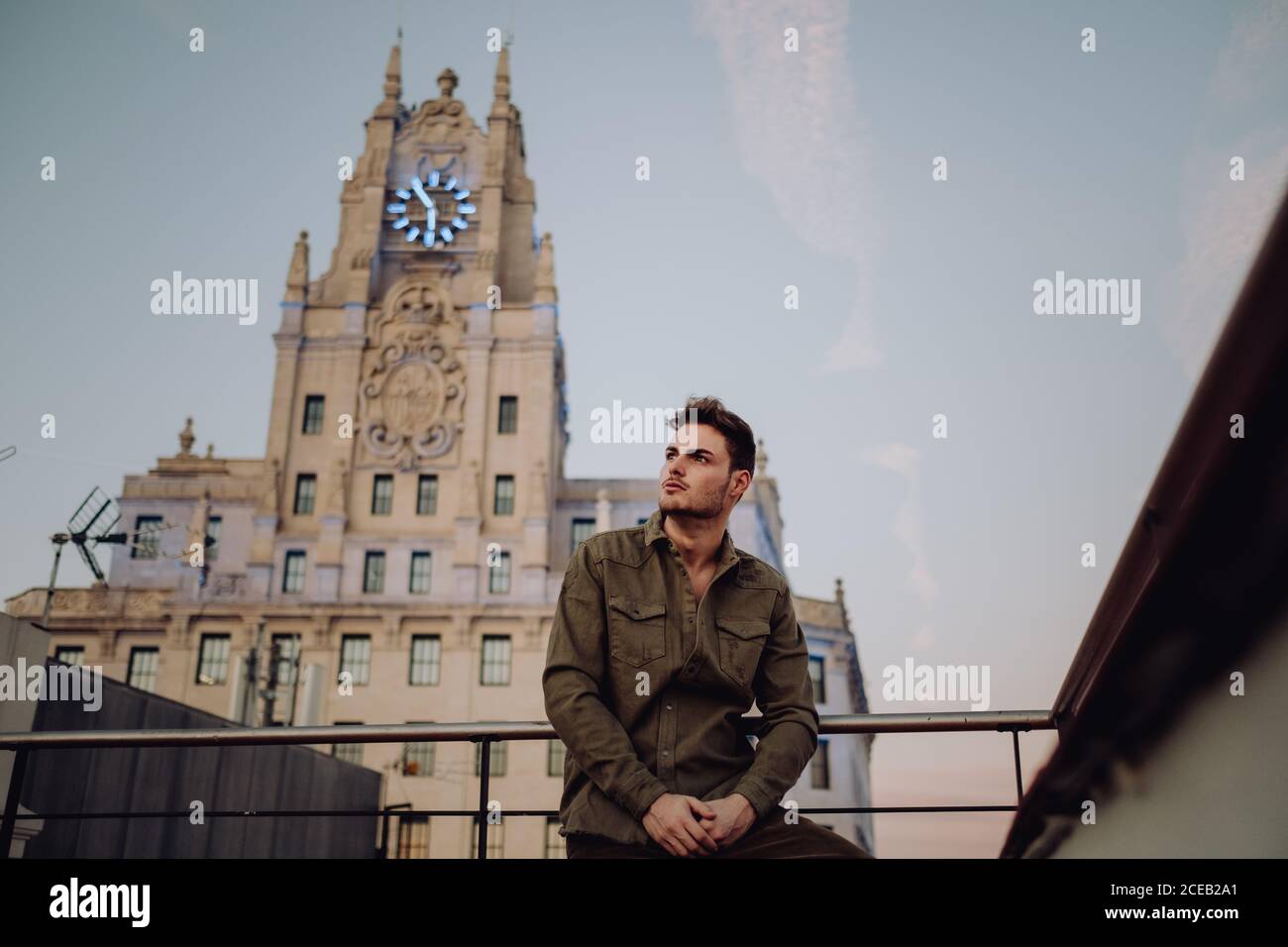 Giovane ragazzo di stile in abbigliamento casual vicino alle rotaie sul tetto che guarda lontano vicino alla vecchia torre con orologi e cielo blu a Madrid, Spagna Foto Stock