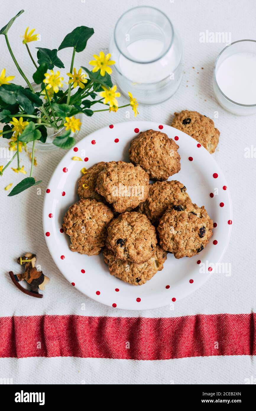 Da sopra la piastra con gustosi biscotti di farina di avena e di vetro e una bottiglia di latte fresco collocato sul tavolo vicino a vaso con graziosi fiori gialli Foto Stock