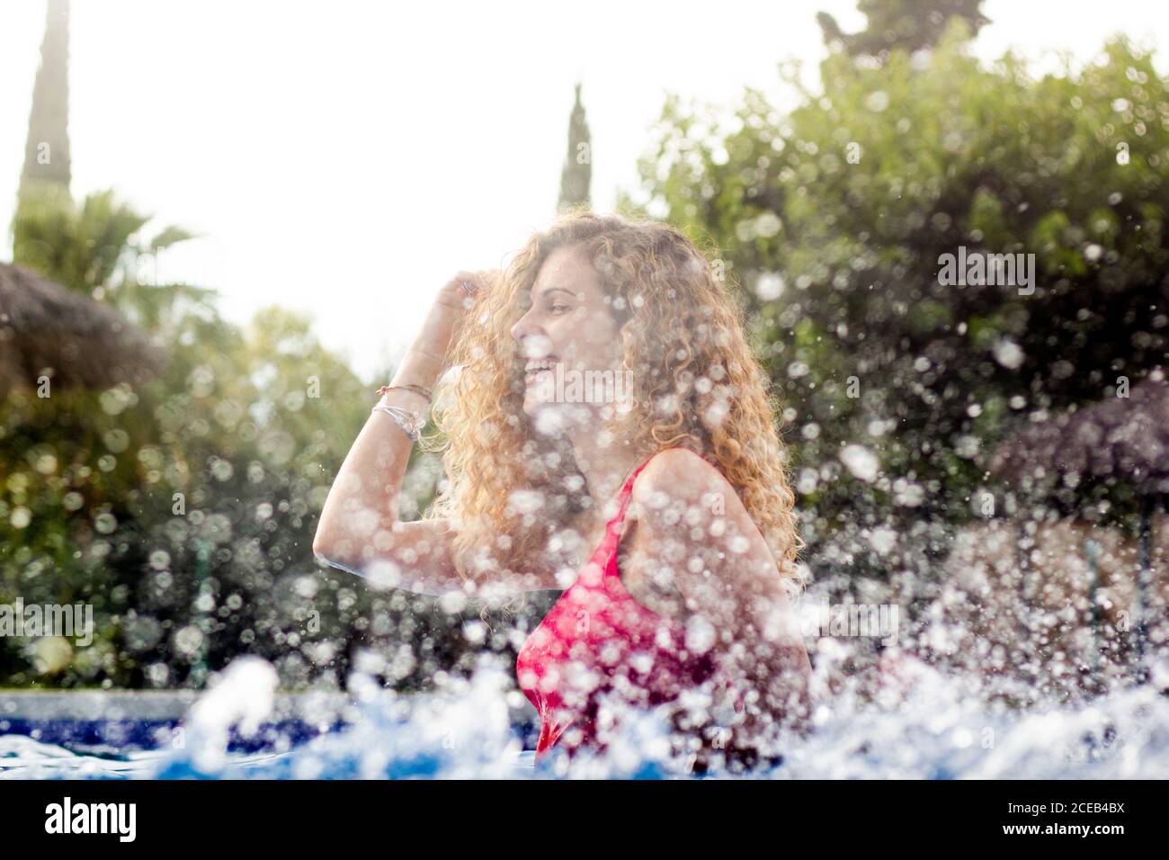 Vista laterale di ricci arrossati Donna regolazione capelli e in piedi in piscina con spruzzi Foto Stock