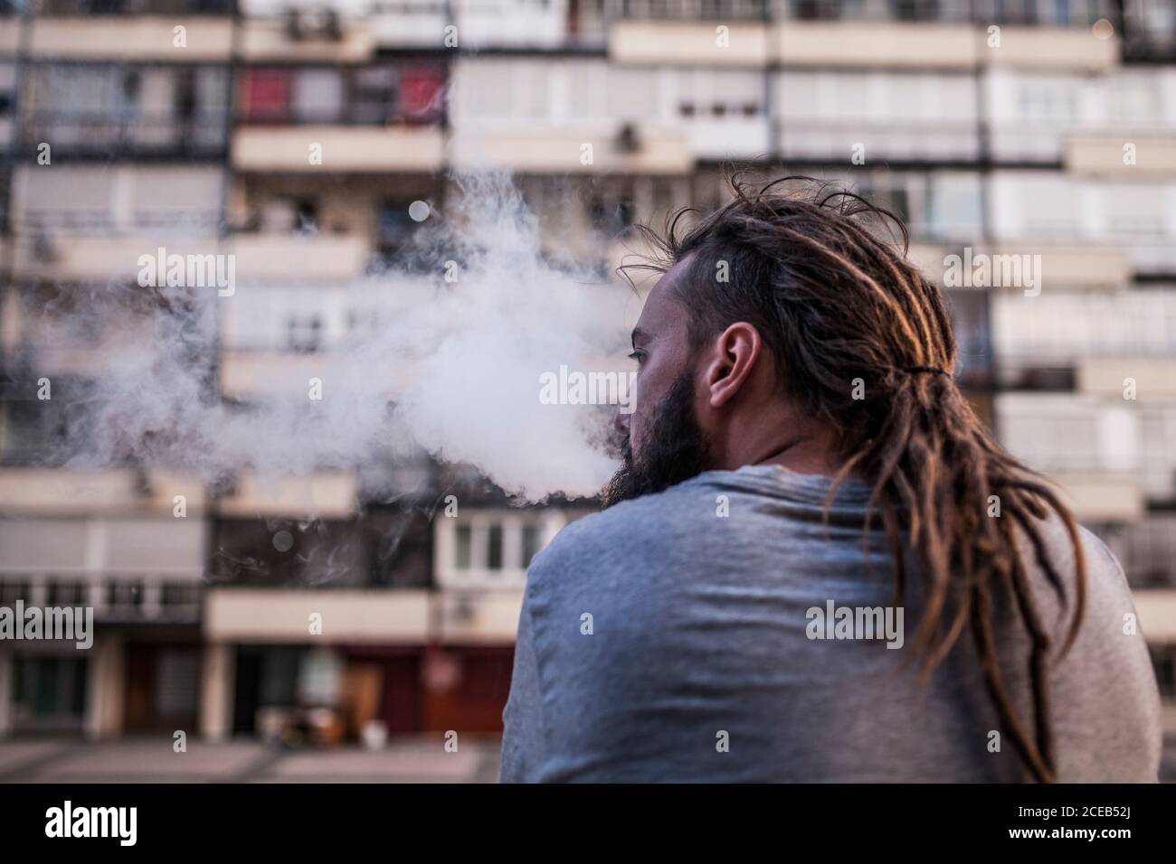 Giovane e caucasico uomo Rastafariano fumare ed esporre il fumo da dietro e guardare a sinistra Foto Stock