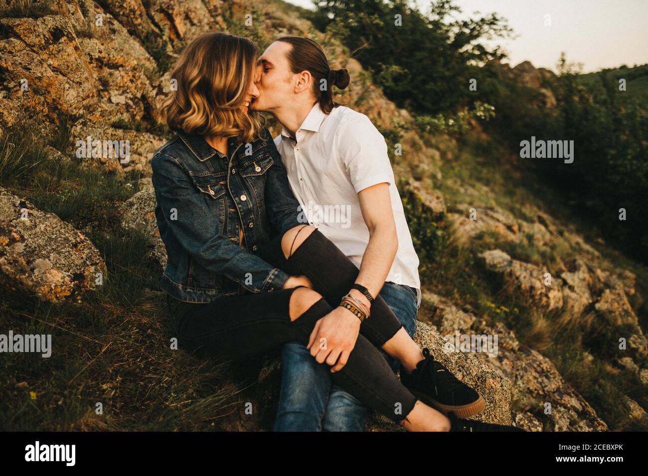 Giovane uomo che bacia la ragazza in guancia mentre si siede insieme pendio di montagna rocciosa Foto Stock