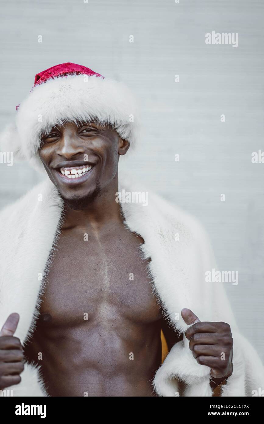 Bell'uomo afro-americano in pelliccia e Babbo Natale cappello sorridente e guardando la macchina fotografica mentre si sta in piedi vicino al bianco parete Foto Stock