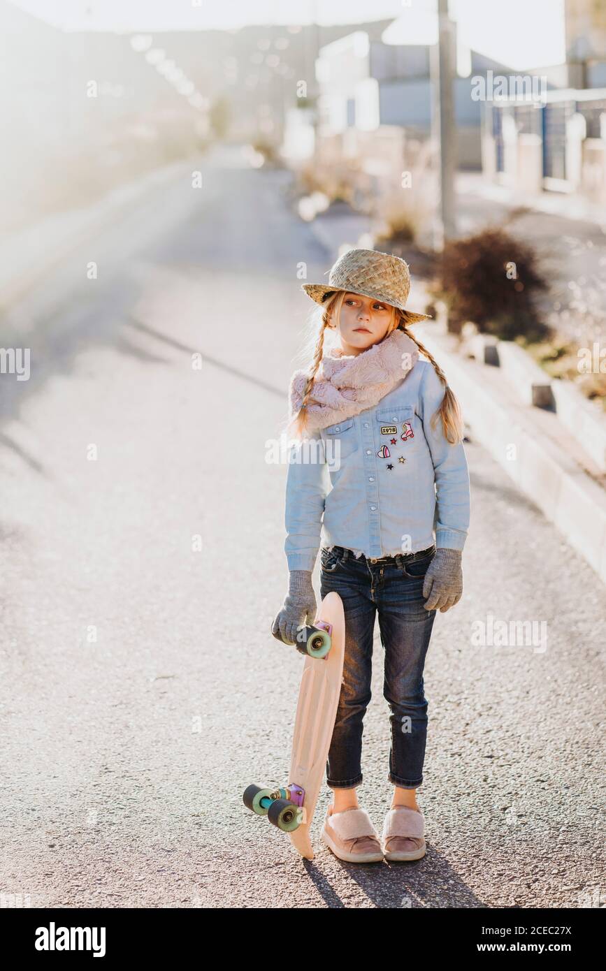 Carino bambina vestito alla moda azienda skateboard e guardando lontano mentre permanente sulla strada sulla giornata di sole sulla strada suburbana Foto Stock