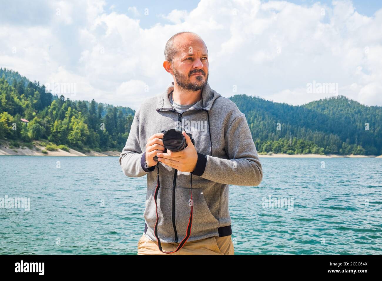 Fotografo uomo in felpa con cappuccio che cammina intorno al lago con fotocamera DSLR e la natura di scatto, ritratto mezzo corpo, concetto di fotografia di paesaggio Foto Stock