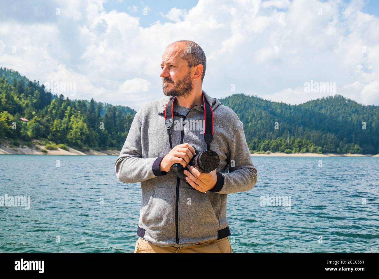 Fotografo uomo in felpa con cappuccio che cammina intorno al lago con fotocamera DSLR e la natura di scatto, ritratto mezzo corpo, concetto di fotografia di paesaggio Foto Stock