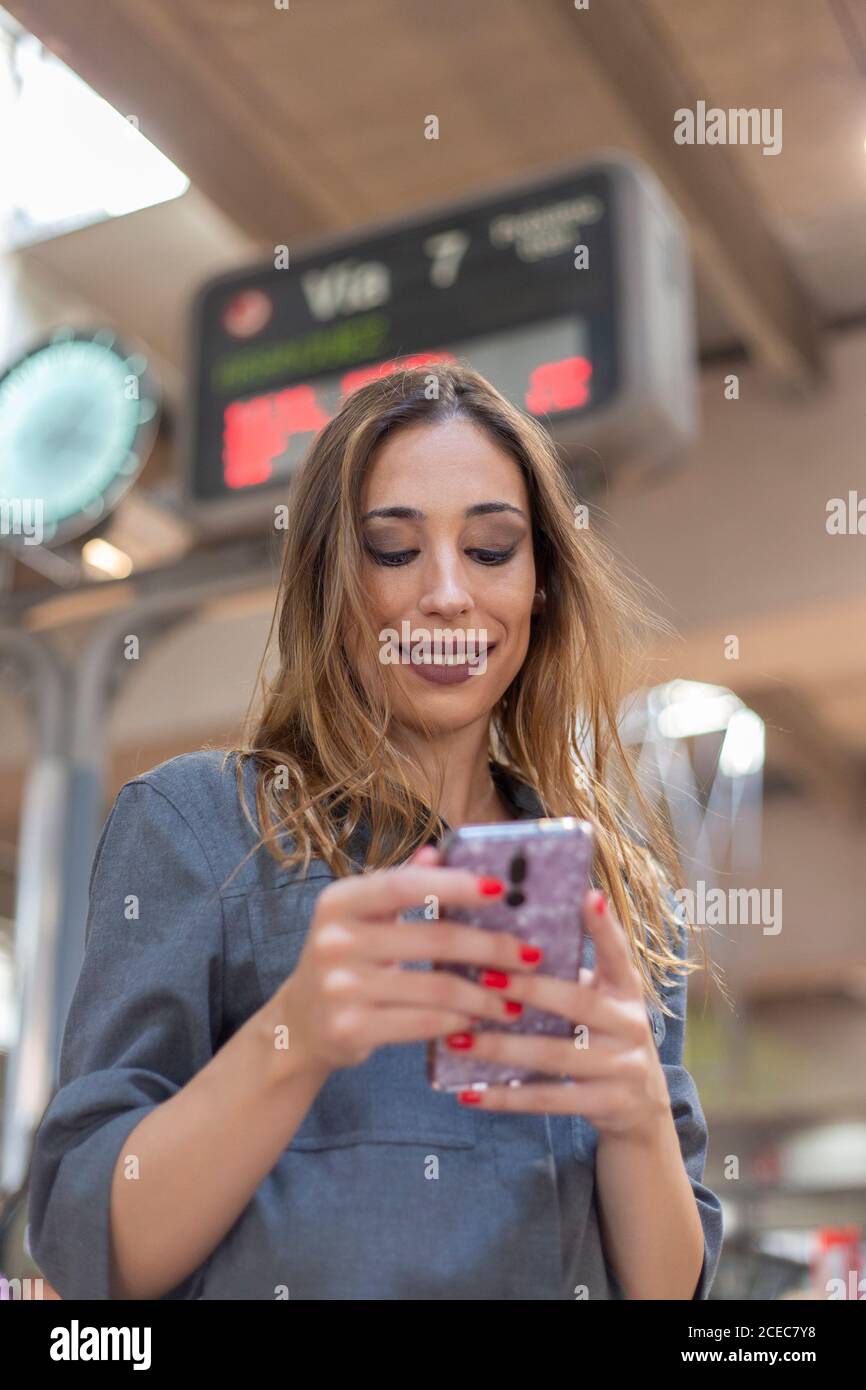 Da sotto colpo di attraente giovane femmina sorridente e di navigazione smartphone moderno mentre ci si trova su uno sfondo sfocato della stazione ferroviaria Foto Stock