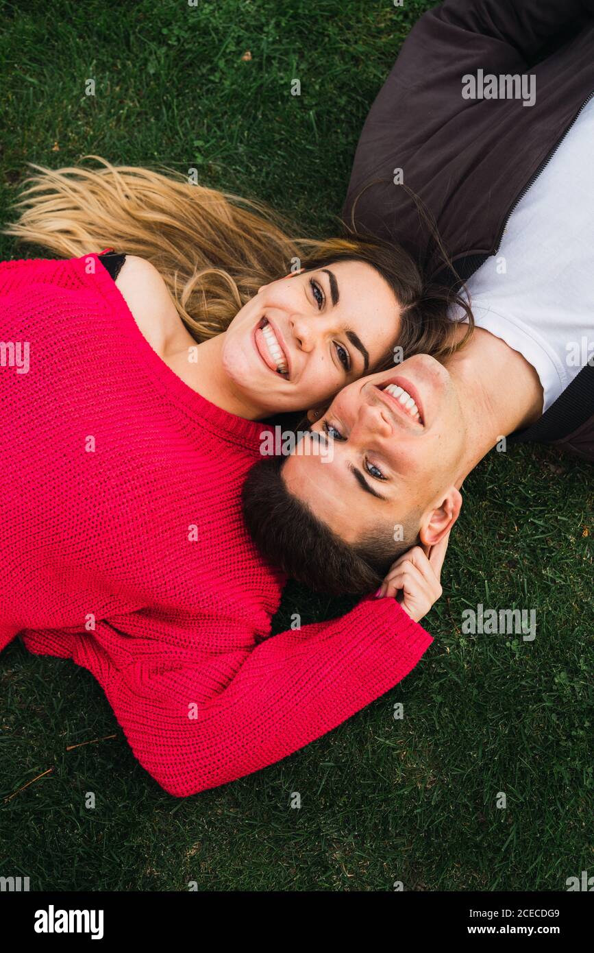 Dall'alto di una coppia allegra e trendy che giace sull'erba verde e guarda la macchina fotografica. Foto Stock
