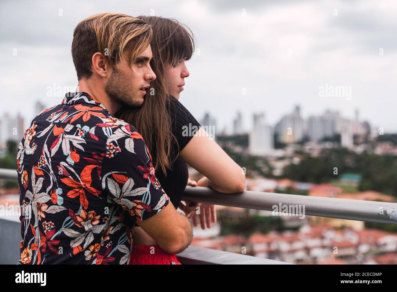 Vista laterale di un giovane ragazzo in un elegante vestito che abbraccia una bella donna dal retro mentre si alza sul balcone e si ammira la vista della città moderna insieme Foto Stock