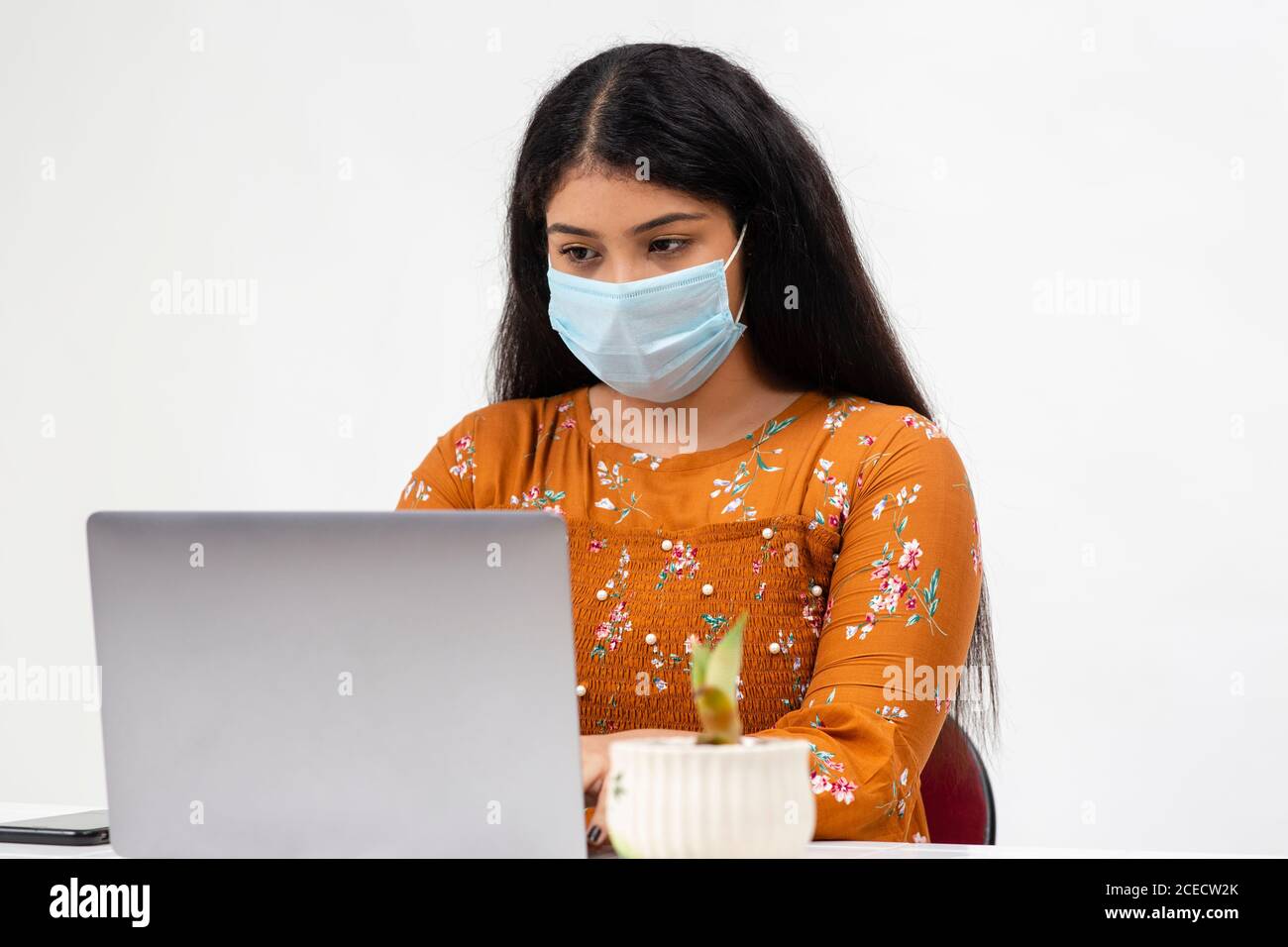 Bella giovane donna indiana seduta su sfondo bianco e utilizzando il suo laptop. Adolescente carino che fa il suo lavoro scolastico in linea da casa. Foto Stock