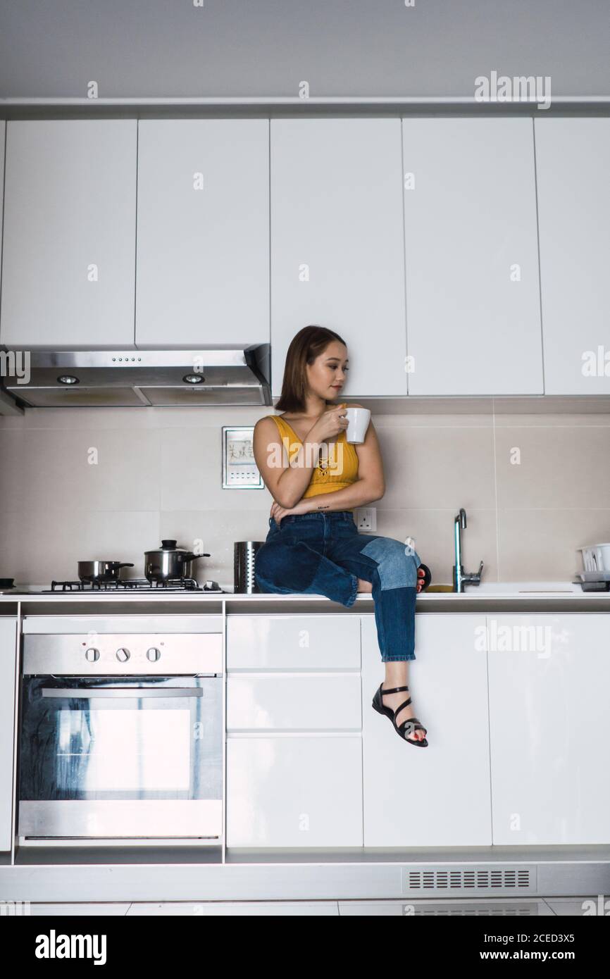 Bella giovane donna asiatica seduta e rilassante con tazza sul tavolo da cucina a casa. Foto Stock
