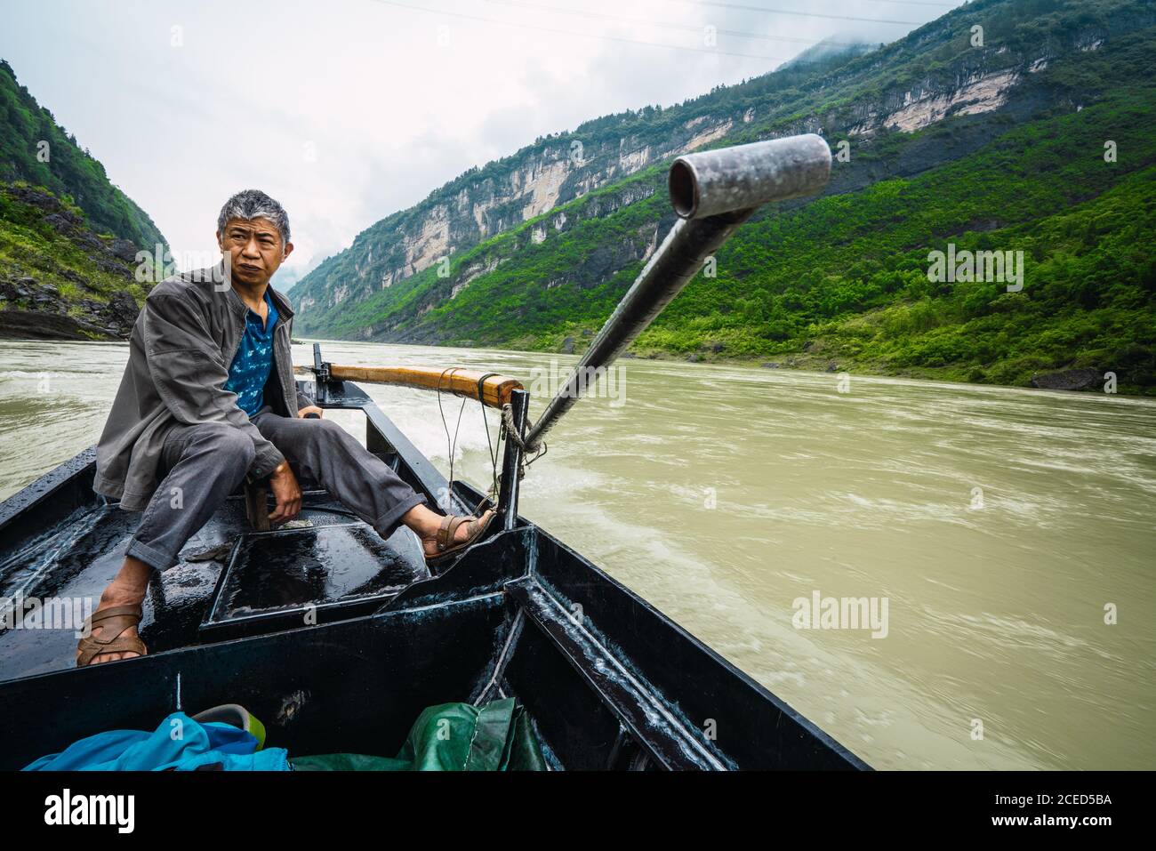 FIUME YANGTZE, CINA - 14 GIUGNO 2018: Uomo cinese anziano guardando la macchina fotografica seduto sulla barca da pesca che galleggia lungo il più lungo in Cina e Asia Yangtze fiume con alte montagne su entrambi i lati e bellissimo cielo sereno sopra Foto Stock