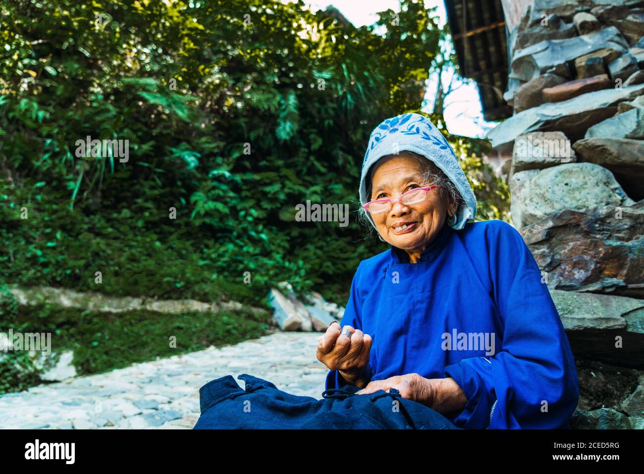 GUINZHOU, CINA - 14 GIUGNO 2018: Vista laterale del raccolto di donna cinese anziana in occhiali e abiti tradizionali blu e cappellino cucito in stoffa mani e guardando lontano seduto su pietre con l'altra donna del gruppo etnico Miao e gli alberi su sfondo sfocato a Guizhou, Cina Foto Stock