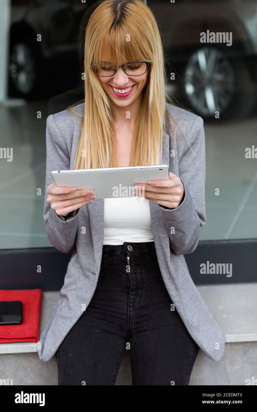 Sorridente giovane signora in occhiali e stile di affari usando lei dispositivo bianco con auto sullo sfondo Foto Stock