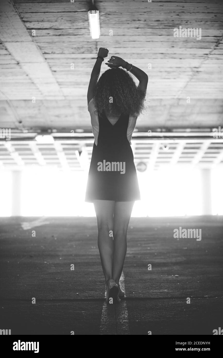 Vista posteriore di Slim Woman con ricci indossando abito nero con sneakers camminare nel parcheggio sotterraneo alla luce del sole Foto Stock