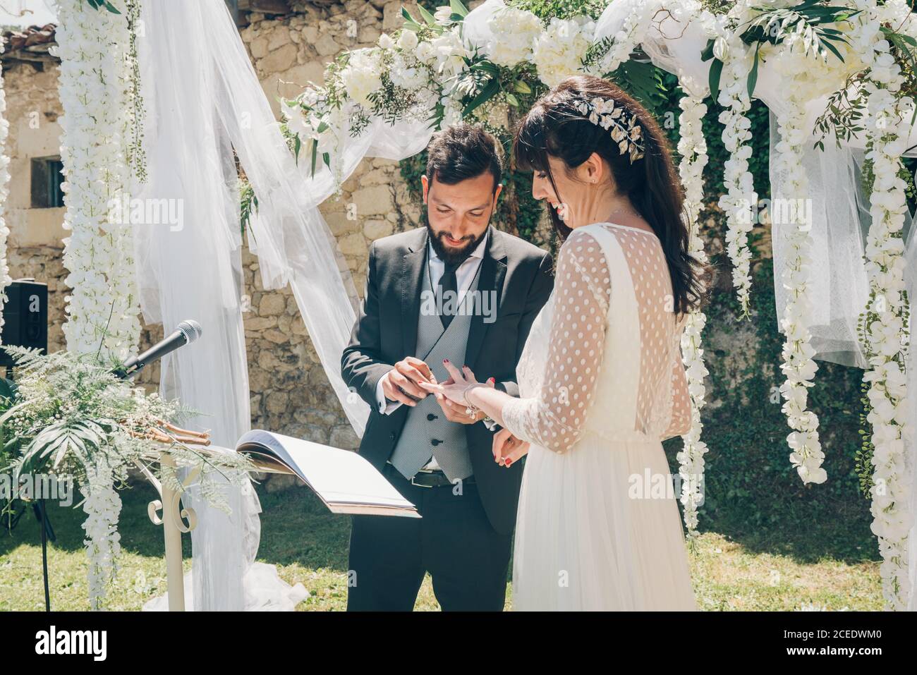 Giovane bella sposa in abito bianco e bello sposo in costume nero che si scambiano anelli su sfondo di decorazioni di nozze Foto Stock