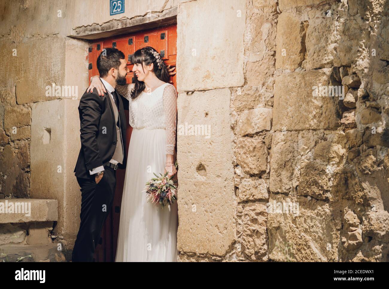 Felice bella sposa in abito da sposa che tiene bouquet e sposo in costume nero sorridendo e abbracciandosi vicino alla pietra parete Foto Stock