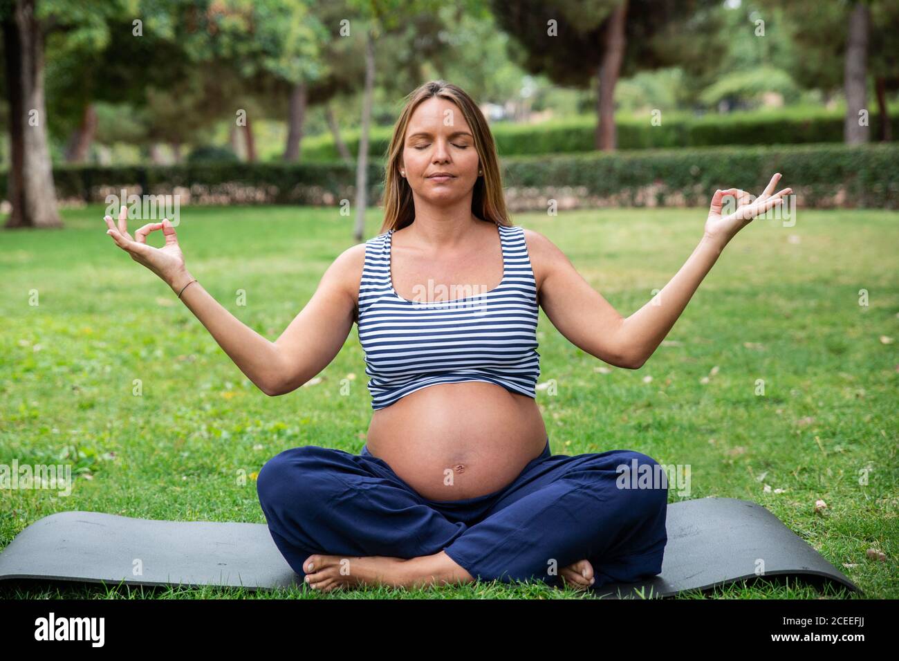 Giovane donna incinta in sport che medita in lotus posa su tappetino yoga in giardino verde Foto Stock