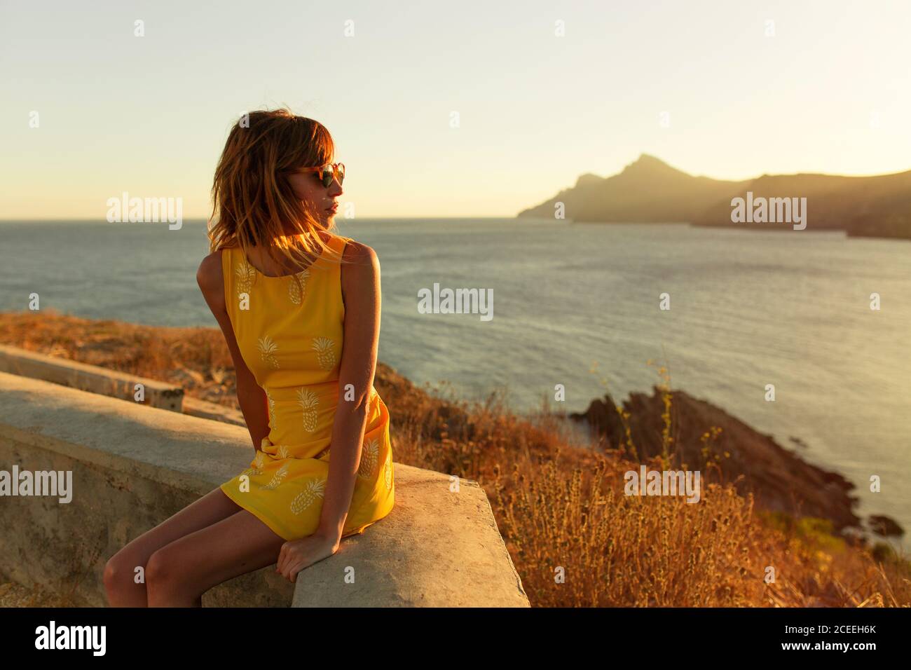 Bella donna in elegante abito corto e occhiali da sole seduti in terrazza con una splendida vista sul mare al tramonto Foto Stock