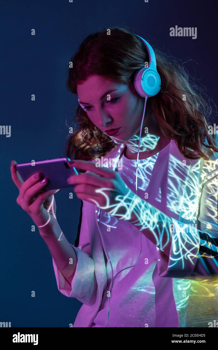 Giovane donna che guarda lo smartphone e indossa le cuffie al neon in studio Foto Stock
