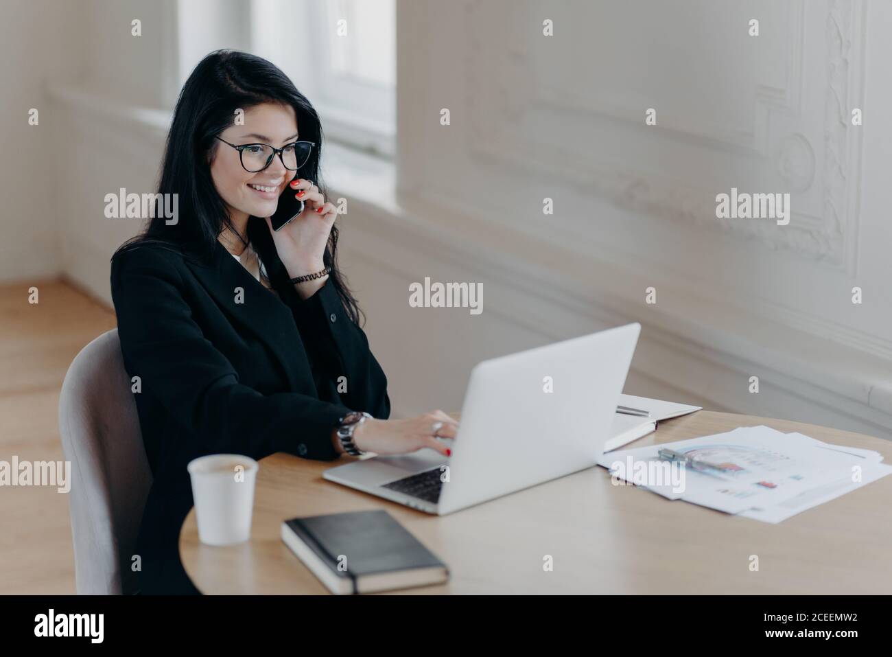 Bella dirigente femminile rende lavoro remoto, parla via telefono cellulare e controlla le informazioni in computer portatile posa sul luogo di lavoro circondato da caffè Foto Stock