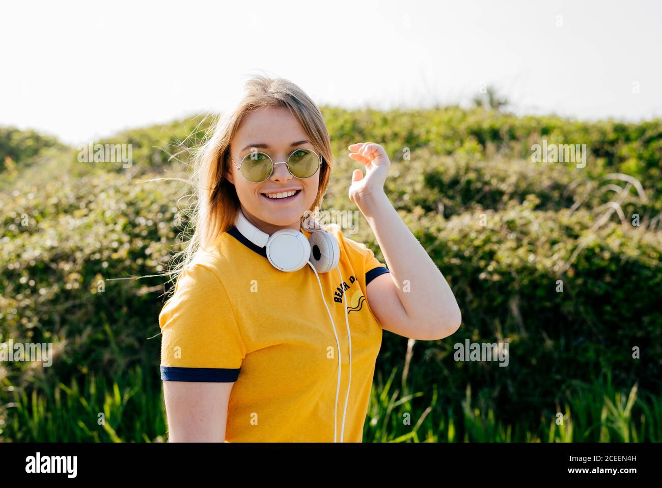 Giovane ragazza casual che indossa occhiali da sole con t-shirt gialla e cuffie sul collo sorridendo alla macchina fotografica nella natura verde. Foto Stock