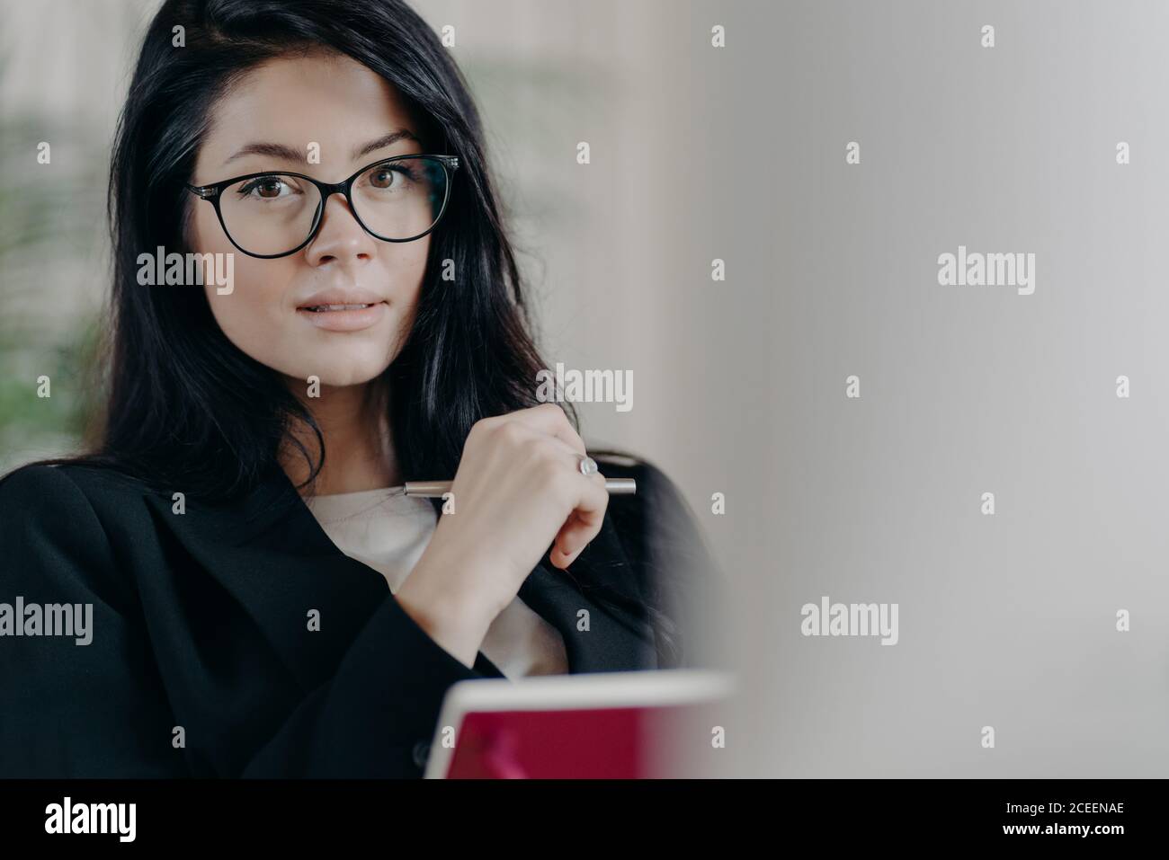 Dipendente donna adulto serio sicuro di sé guarda anche se occhiali trasparenti, scrive informazioni mentre lavora sul progetto, fa piani, vestito in fo Foto Stock