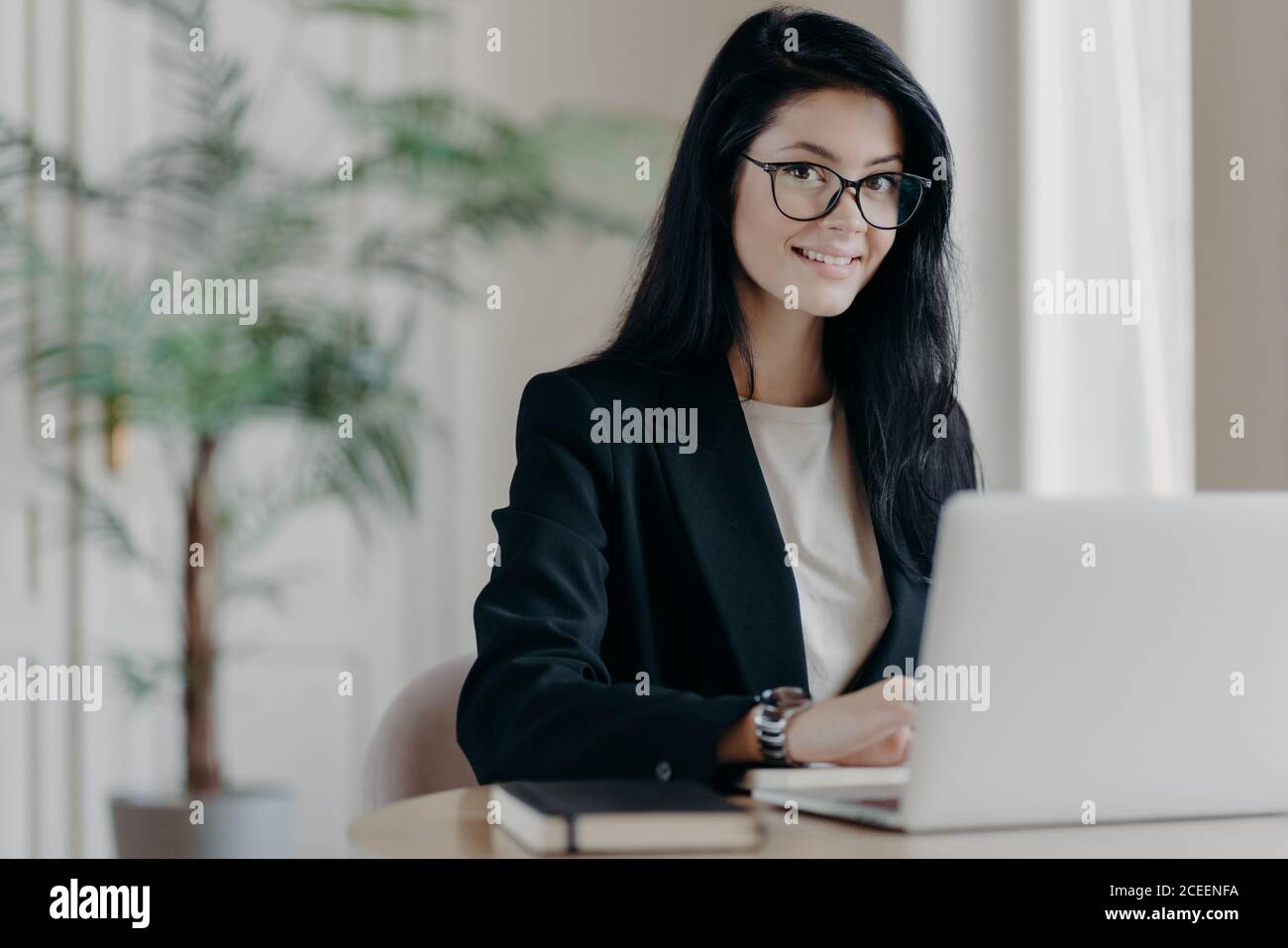 Sorridente giovane donna ben vestito segretaria con capelli scuri, lavora su computer portatile a desktop, fa progetto o lavoro di ricerca. Responsabile vendite in moder Foto Stock