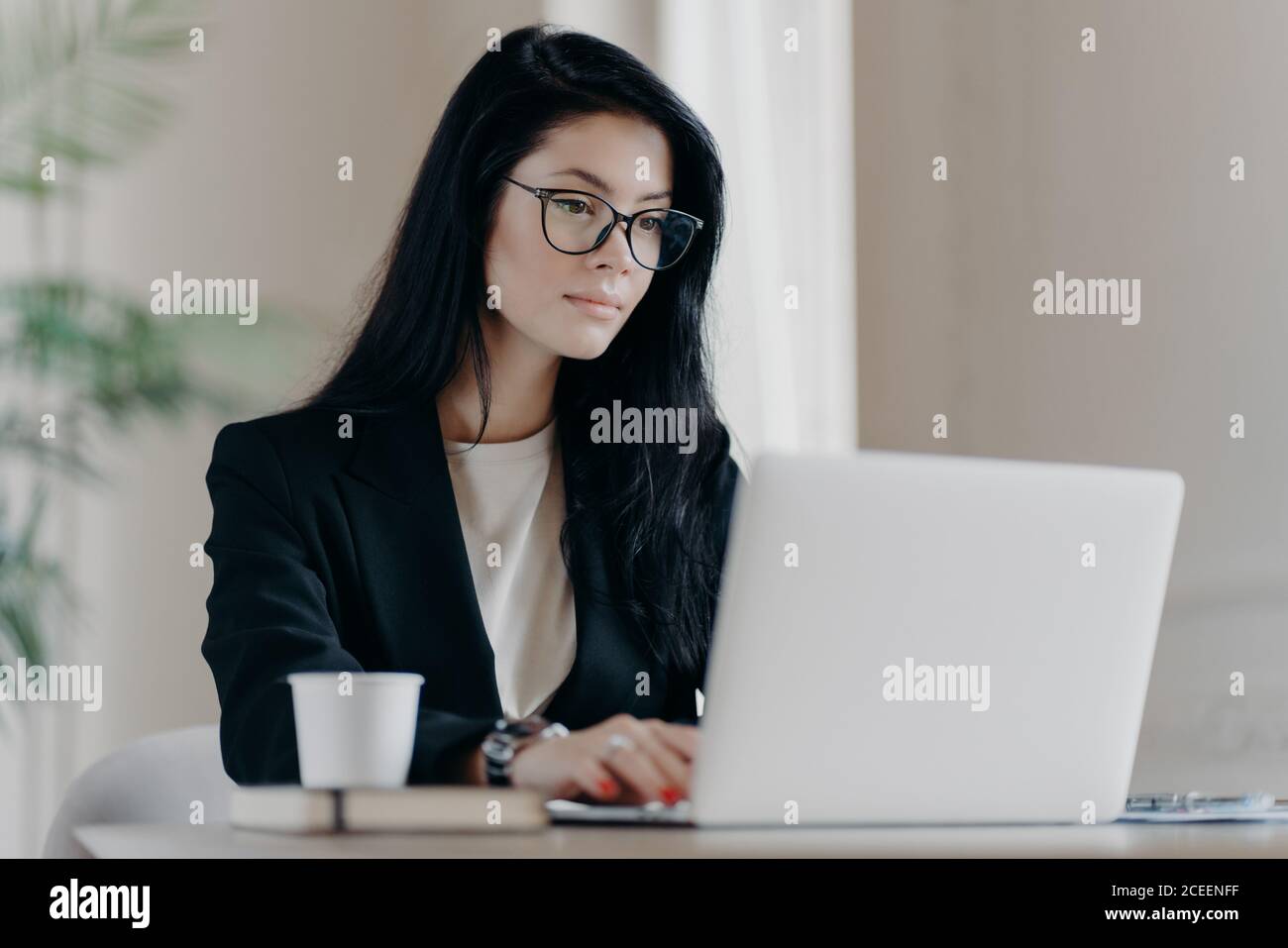 Concentrato donna brunette specialista IN occhiali ricerca informazioni utilizzando internet, tastiere su computer portatile, bevande caffè, ha gravi exp Foto Stock