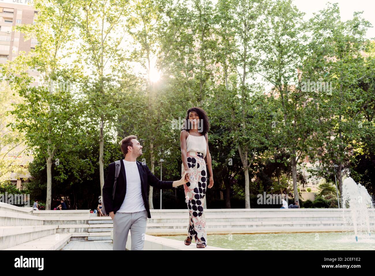 Affascinante donna nera sorridendo e camminando sul bordo della fontana vicino al ragazzo mentre si passa il tempo nel parco insieme Foto Stock