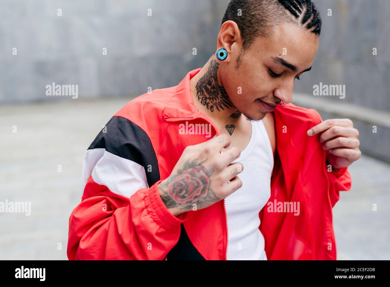 Hipster sorridente con piercing e tunnel in orecchie che tengono rosso giacca casual in mani su sfondo grigio sfocato Foto Stock