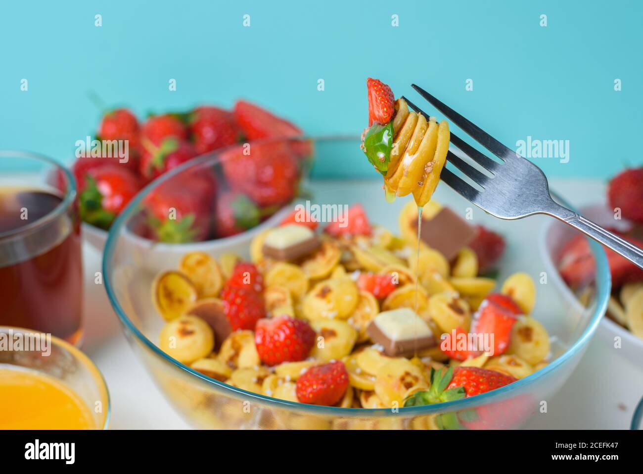 minuscoli pancake su una forchetta con fragole fresche, banana, cioccolato, patatine di cocco e miele in un piatto su sfondo blu. tendenza alimentare 2020. Foto Stock