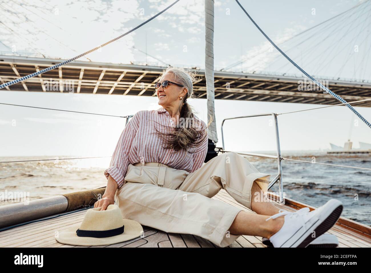 Donna anziana di stile con i capelli lunghi che si guardano via mentre si siede su un arco di yacht. Ragazza matura che si gode di viaggio in barca a vela. Foto Stock