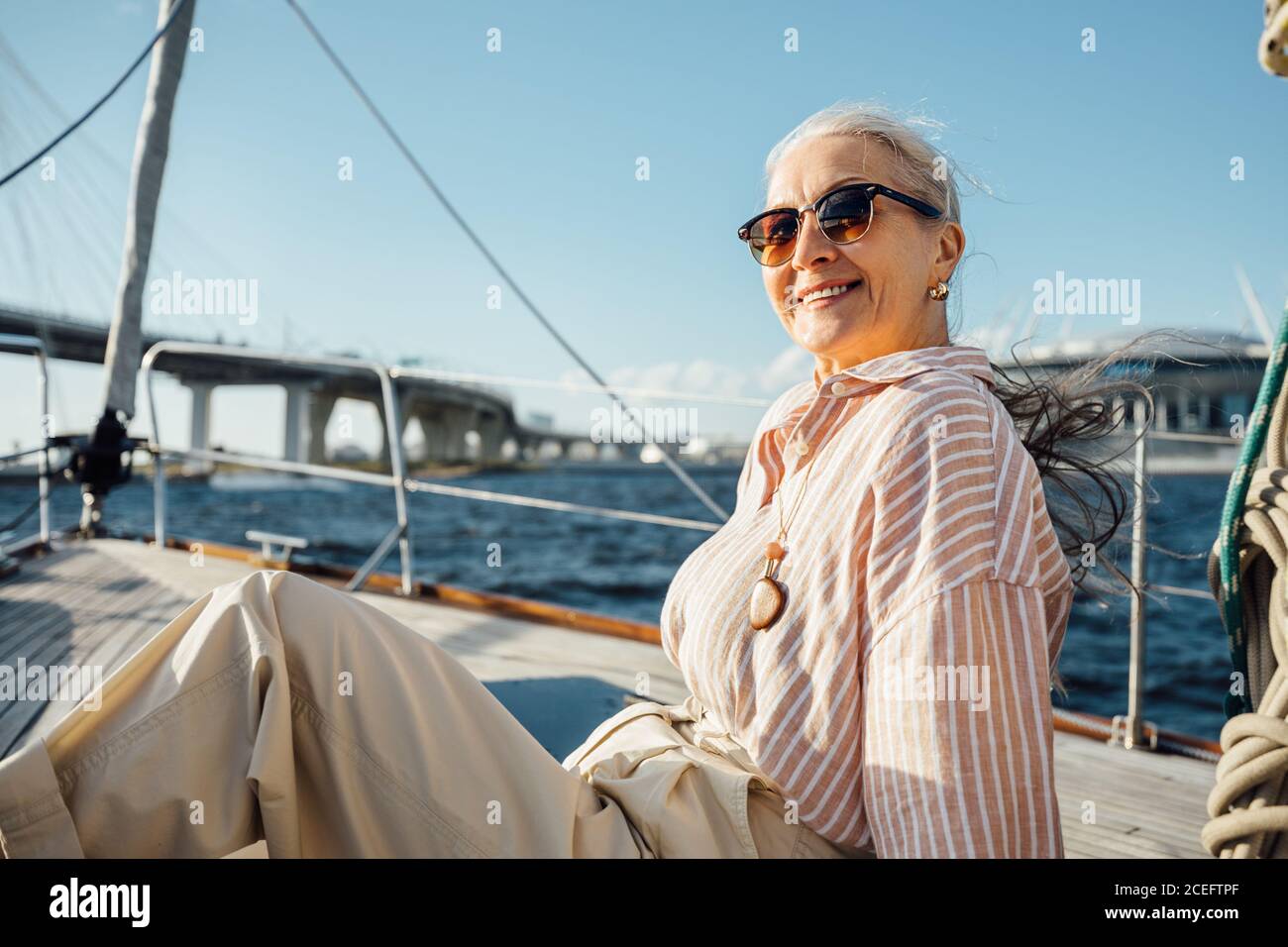 Elegante donna matura che indossa occhiali su uno yacht privato e guarda direttamente una macchina fotografica. Donna sorridente seduta su una barca a vela e godendosi una vacanza Foto Stock
