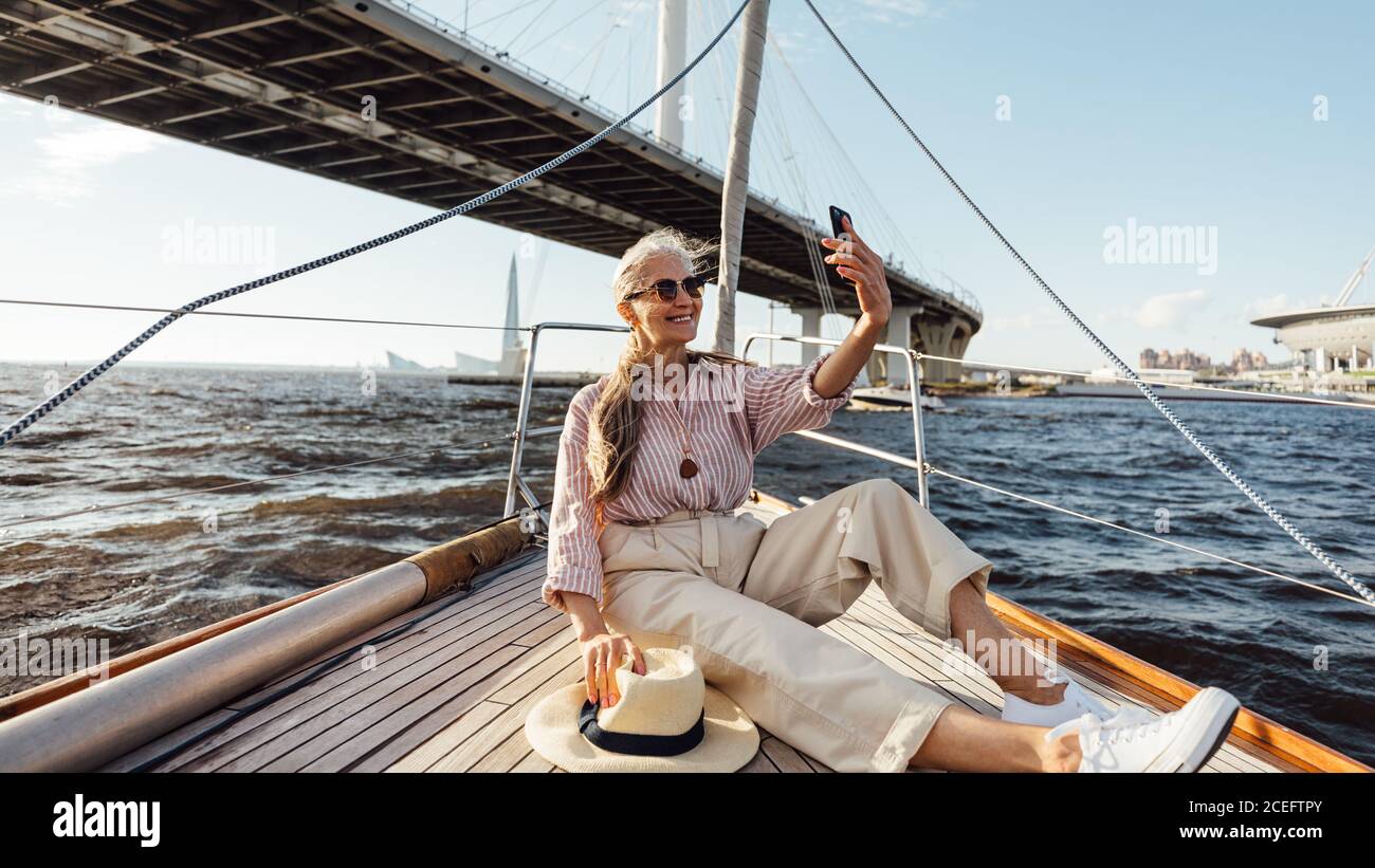 Felice donna matura in yacht viaggio prendendo un selfie. Elegante donna seduta su un ponte di barca scattando fotografie sul telefono cellulare mentre galleggia sotto una sposa Foto Stock
