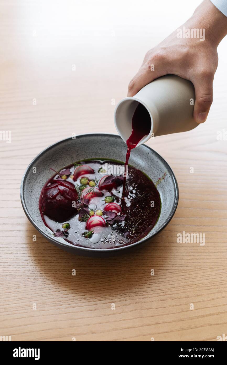 Vista del raccolto della mano maschile che versa succo di barbabietola rosso scuro Dalla caraffa alla ciotola grigia con verdure nordiche tritate e. potherbs posto su tavolo di legno chiaro Foto Stock