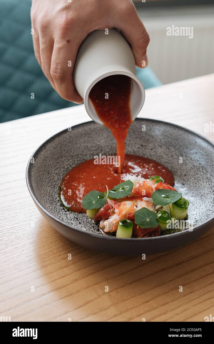 Vista di raccolto della mano maschio che versa salsa spessa rossa da caraffa nel recipiente con insalata tritata con verdure e pesce servita su tavolo di legno Foto Stock