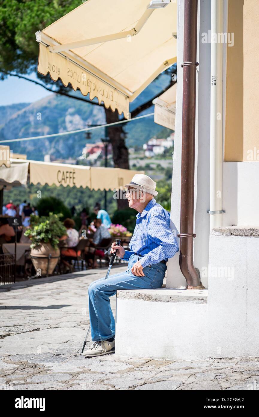Un vecchio uomo italiano locale prende un momento dal sole di metà giornata e guarda la vita passare nella piazza di Ravello, Italia. Foto Stock