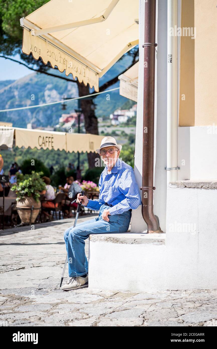 Un vecchio uomo italiano locale prende un momento dal sole di metà giornata e guarda la vita passare nella piazza di Ravello, Italia. Foto Stock
