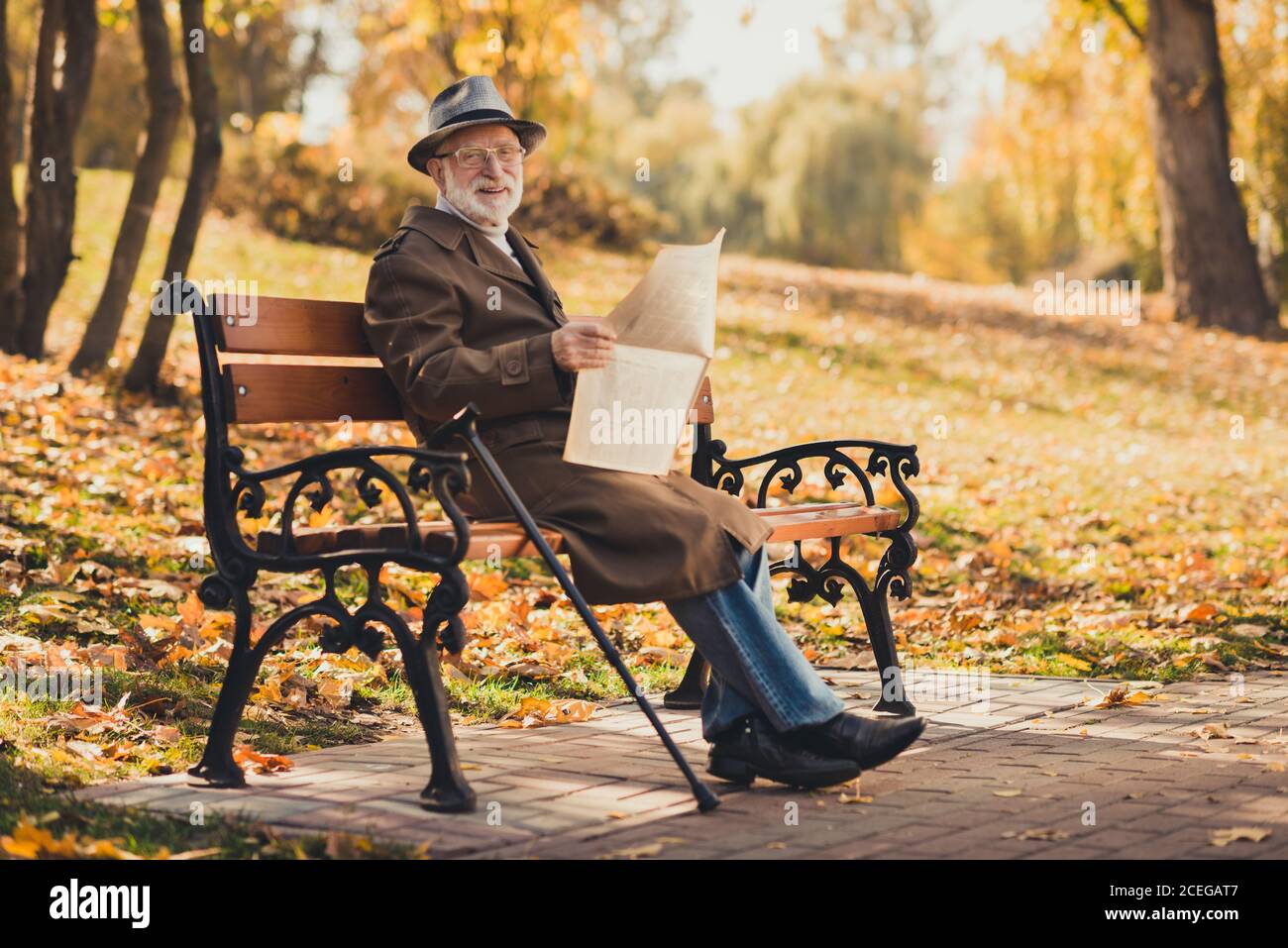 Foto a lunghezza intera di seduta positiva del vecchio uomo seduta panca caduta parco della città fuori sentire amico chiamarlo smettere di leggere il giornale smile indossa un cappellino di stagione Foto Stock