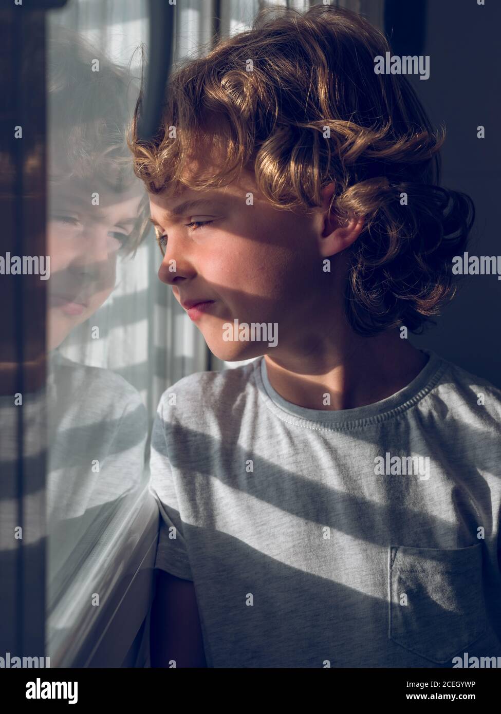 Adorabile ragazzo in T-shirt grigia appoggiata sul vetro e guardando fuori la finestra. Foto Stock