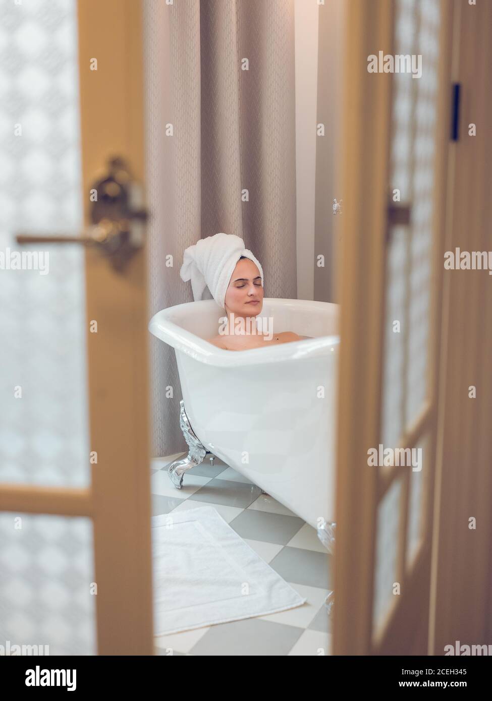 Rilassante bella donna sdraiata nella vasca da bagno con gli occhi chiusi e asciugamano sulla testa visto attraverso le porte aperte a metà. Foto Stock