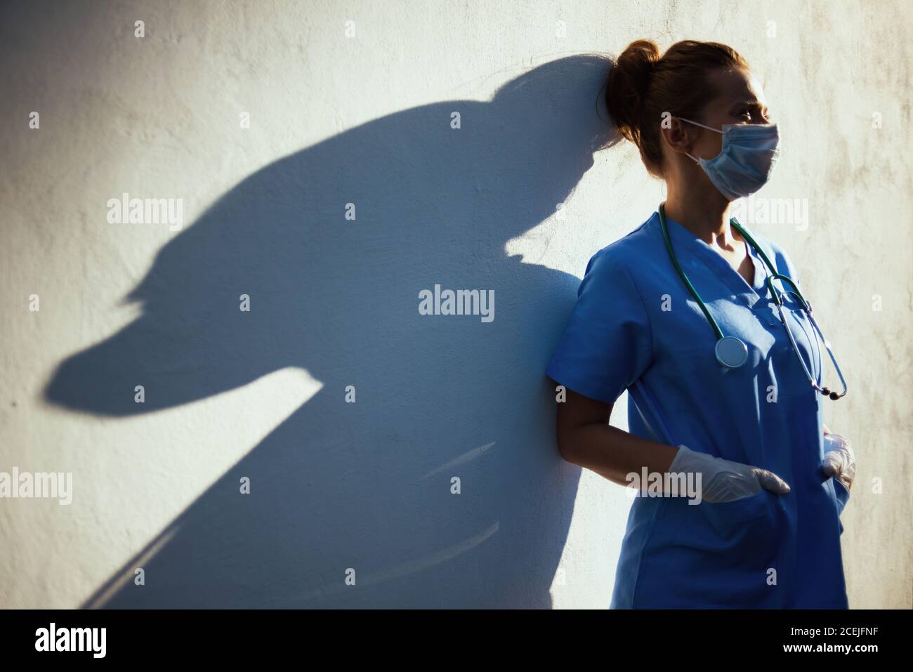 pandemia di coronavirus. donna medica moderna stanca in scrub con stetoscopio, maschera medica e guanti di gomma all'aperto in città vicino a muro. Foto Stock