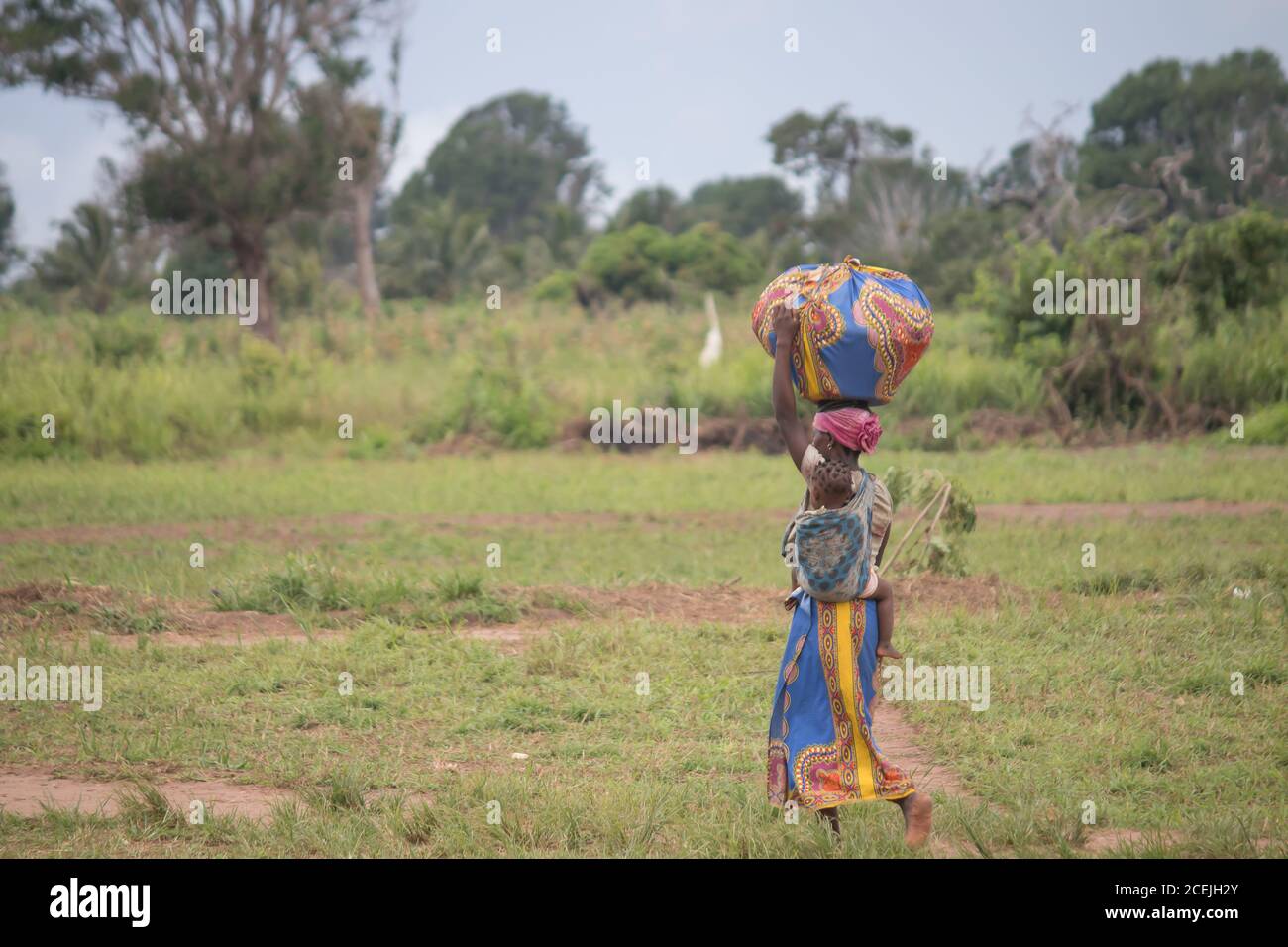 Le donne africane camminano nella natura, vestite con abiti tradizionali colorati, trasportando grandi carichi sulla testa Foto Stock