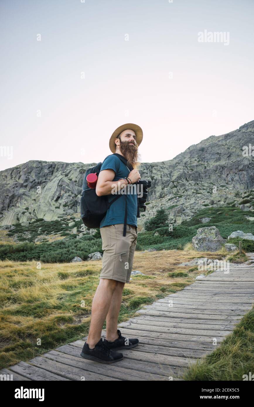 Vista laterale di un giovane bel ragazzo con cappello e barba e zaino in  piedi su strada di legno che conduce alla montagna Foto stock - Alamy