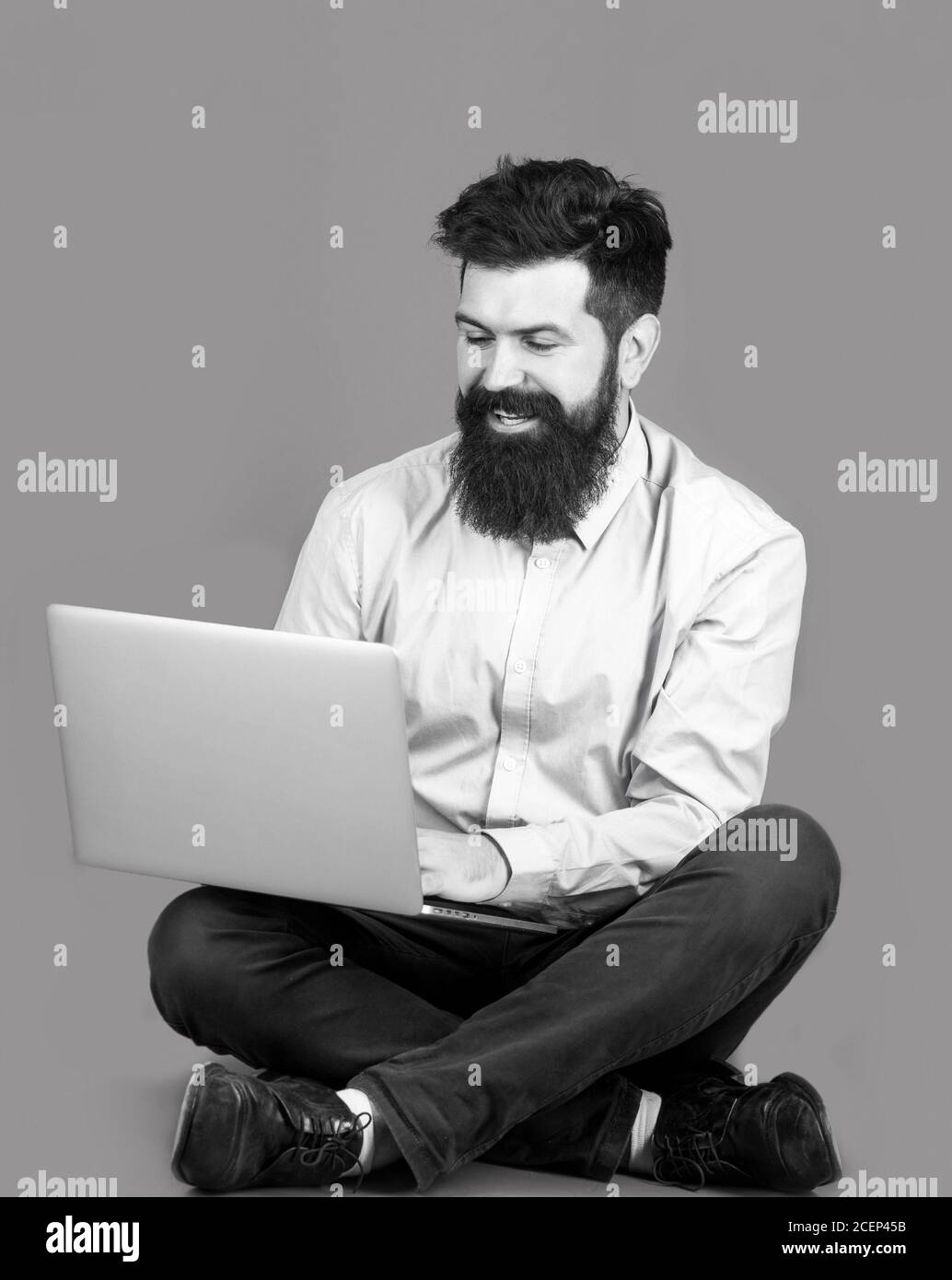 Sorridente bel notebook uomo-operaio con bearded. Felice giovane uomo seduto sul pavimento con e utilizzando il computer portatile su sfondo rosso. Giovane uomo d'affari Foto Stock