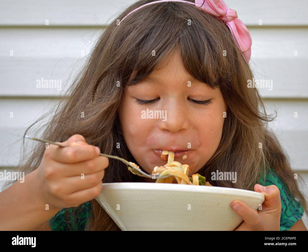 Affamato misto-razza carina bambina (Asia orientale / caucasica) con fiocco rosa nei suoi capelli mangia noodle asciutti con una forchetta da una ciotola di ceramica bianca. Foto Stock