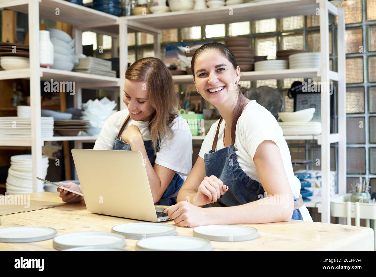 Due donne imprenditrici sorridenti con computer portatile in sala da lavoro artigianale. Vita in su ritratto di allegra donna sorridente artigiana in posa in studio di ceramica e guardando la macchina fotografica Foto Stock